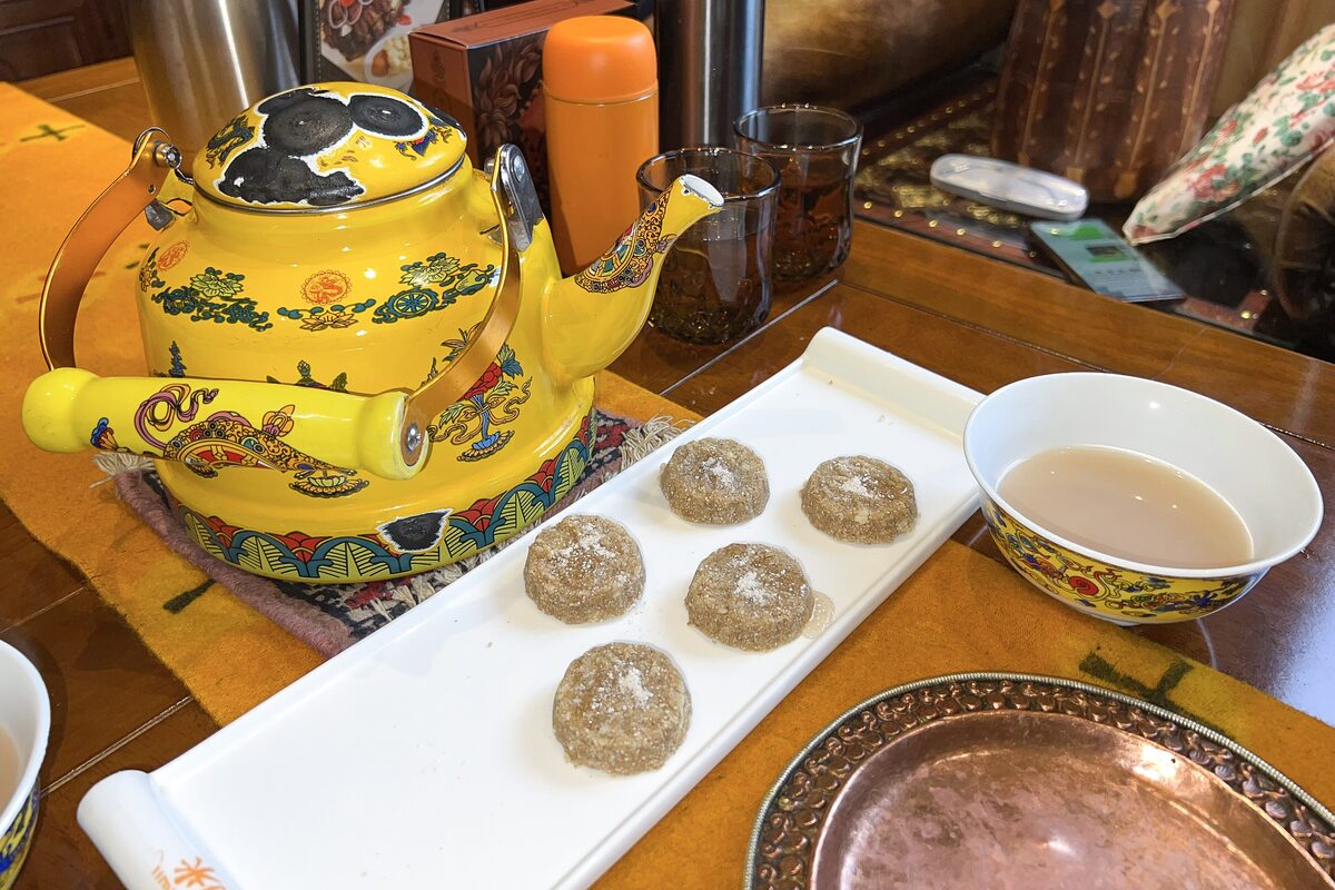 A bowl of butter tea beside a yellow enamel teapot and a plate of small tsampa cakes