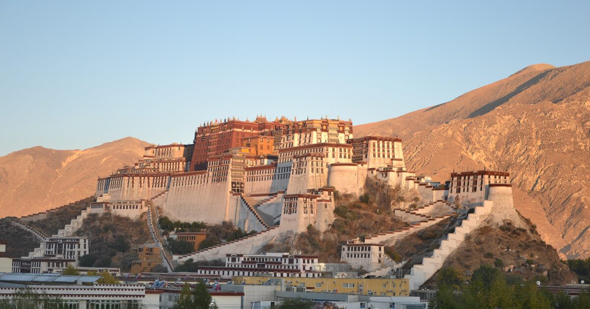 The Potala Palace, white and red walls on its hill in warm low sun under a clear sky, with bare brown mountains behind