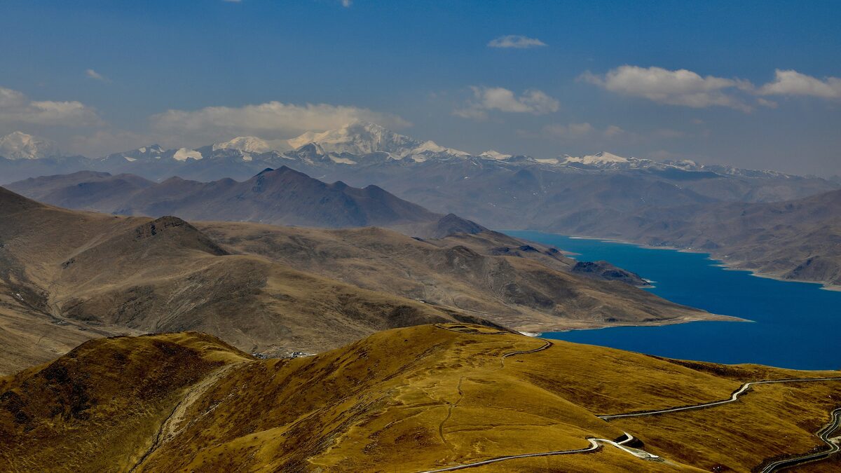 View from the high pass over brown mountain ridges and the blue arm of Yamdrok Lake, with snow peaks on the horizon