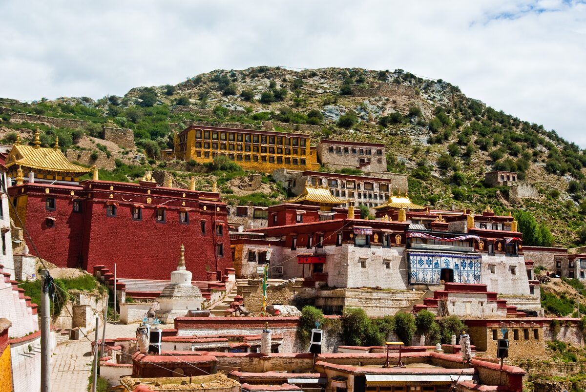 Red and white Ganden Monastery buildings stacked up a green mountainside, with golden roof ornaments