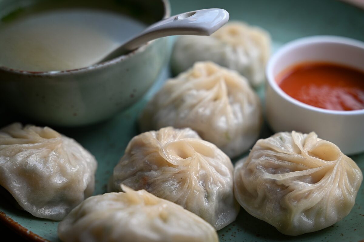 Steamed round momo dumplings with pleated tops on a green plate, next to a bowl of broth and a small dish of red chili dip
