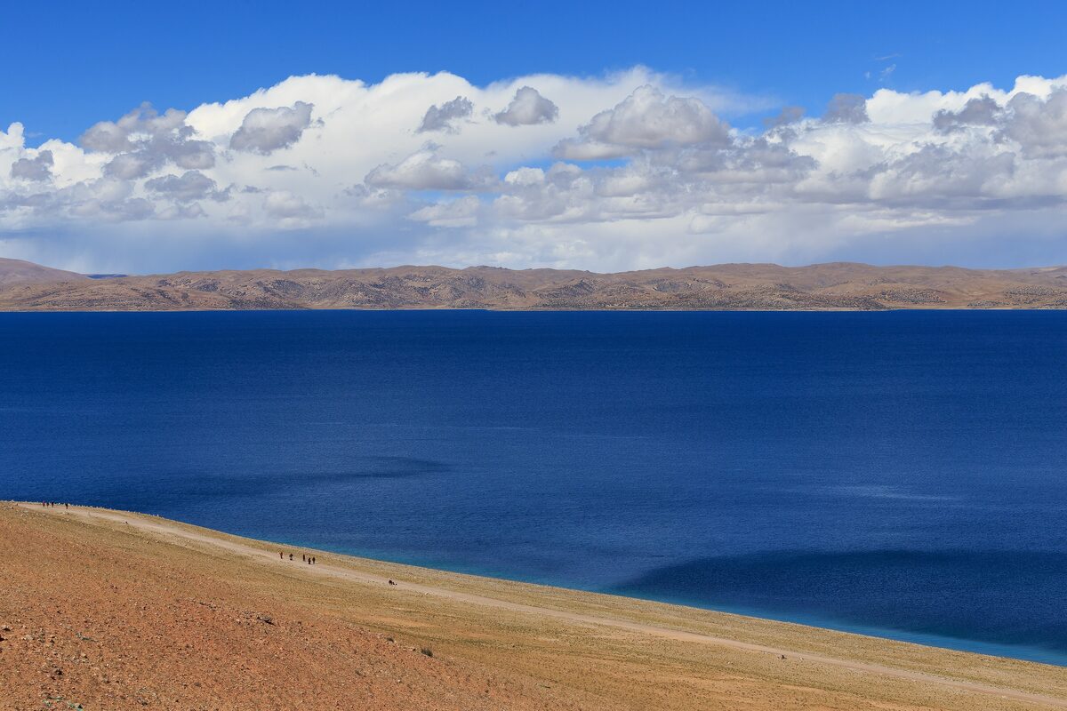 The deep blue water of Namtso under a sky of white clouds, with a low brown mountain ridge on the far shore and bare hillside in the foreground