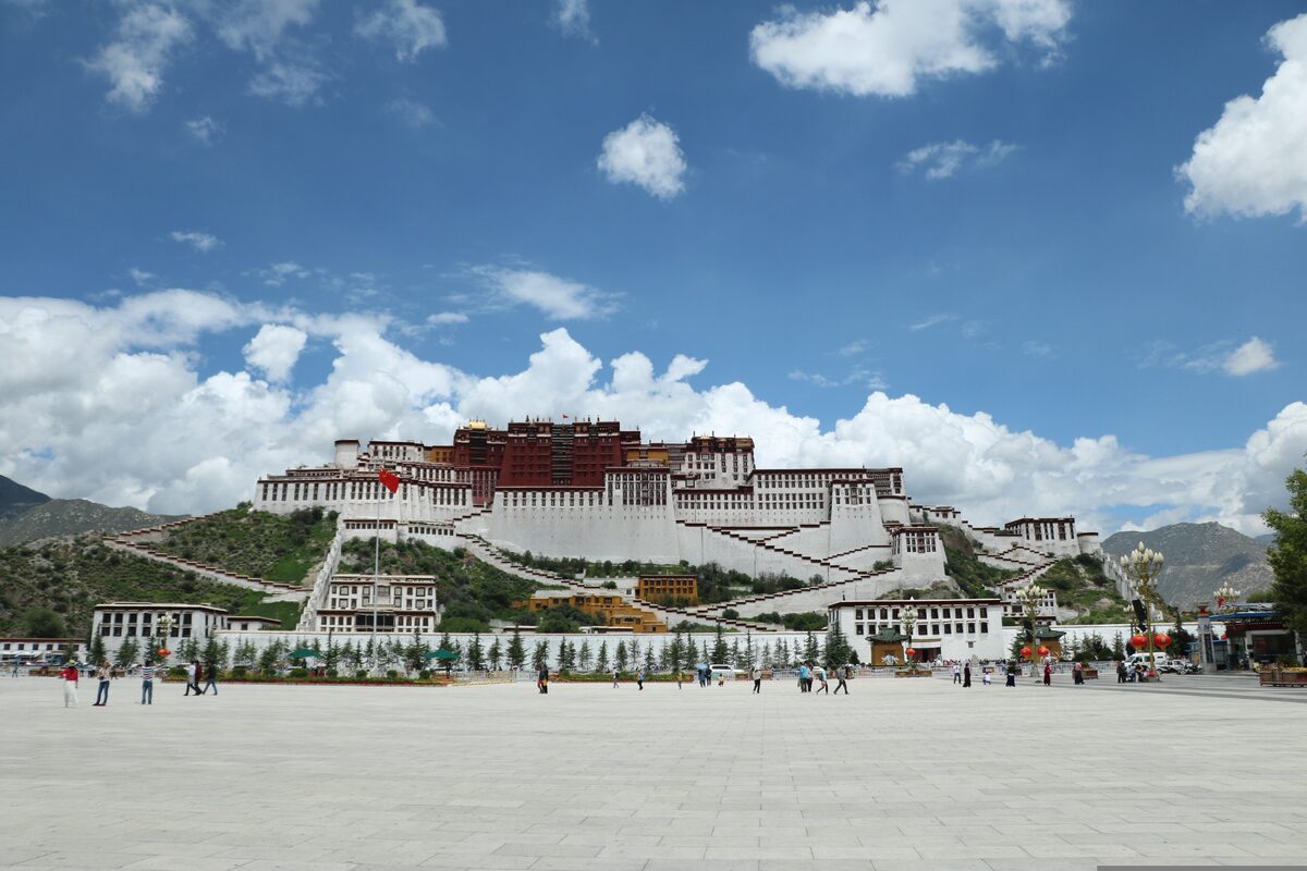 The Potala Palace on its hill across the wide square, white lower walls and the red palace above under blue sky and clouds