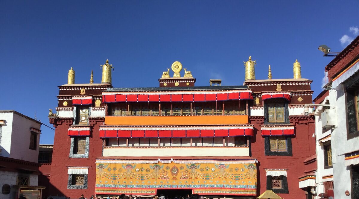 The red and white main hall of Ramoche Temple with golden roof ornaments, red and white awnings and a large embroidered entrance hanging, under deep blue sky