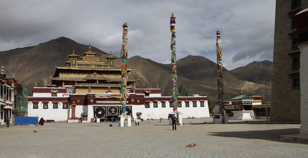 The main hall of Samye Monastery with its golden multi-tiered roof, three painted poles and a wide gravel courtyard before dry hills