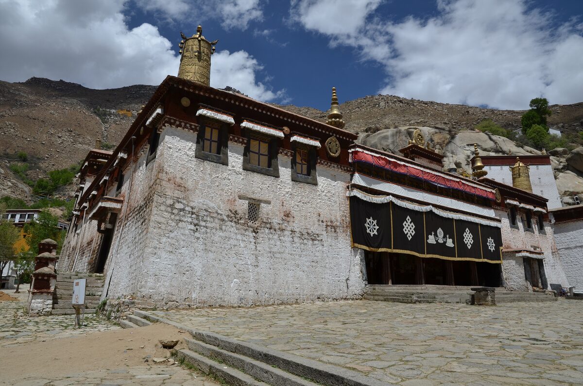 A whitewashed Sera Monastery hall with a black embroidered entrance curtain and a golden roof finial under blue sky