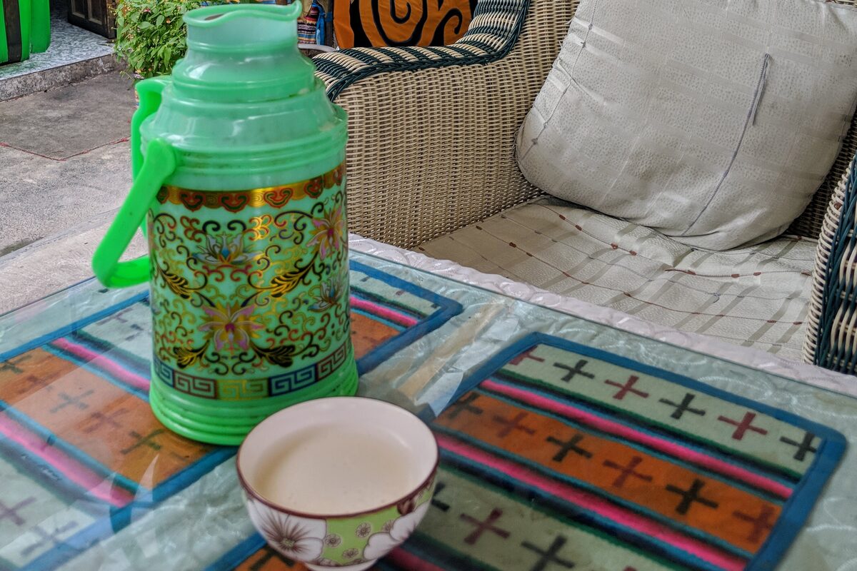 A green patterned thermos beside a small cup of butter tea on a glass-topped table with Tibetan-pattern place mats at a Tibetan teahouse