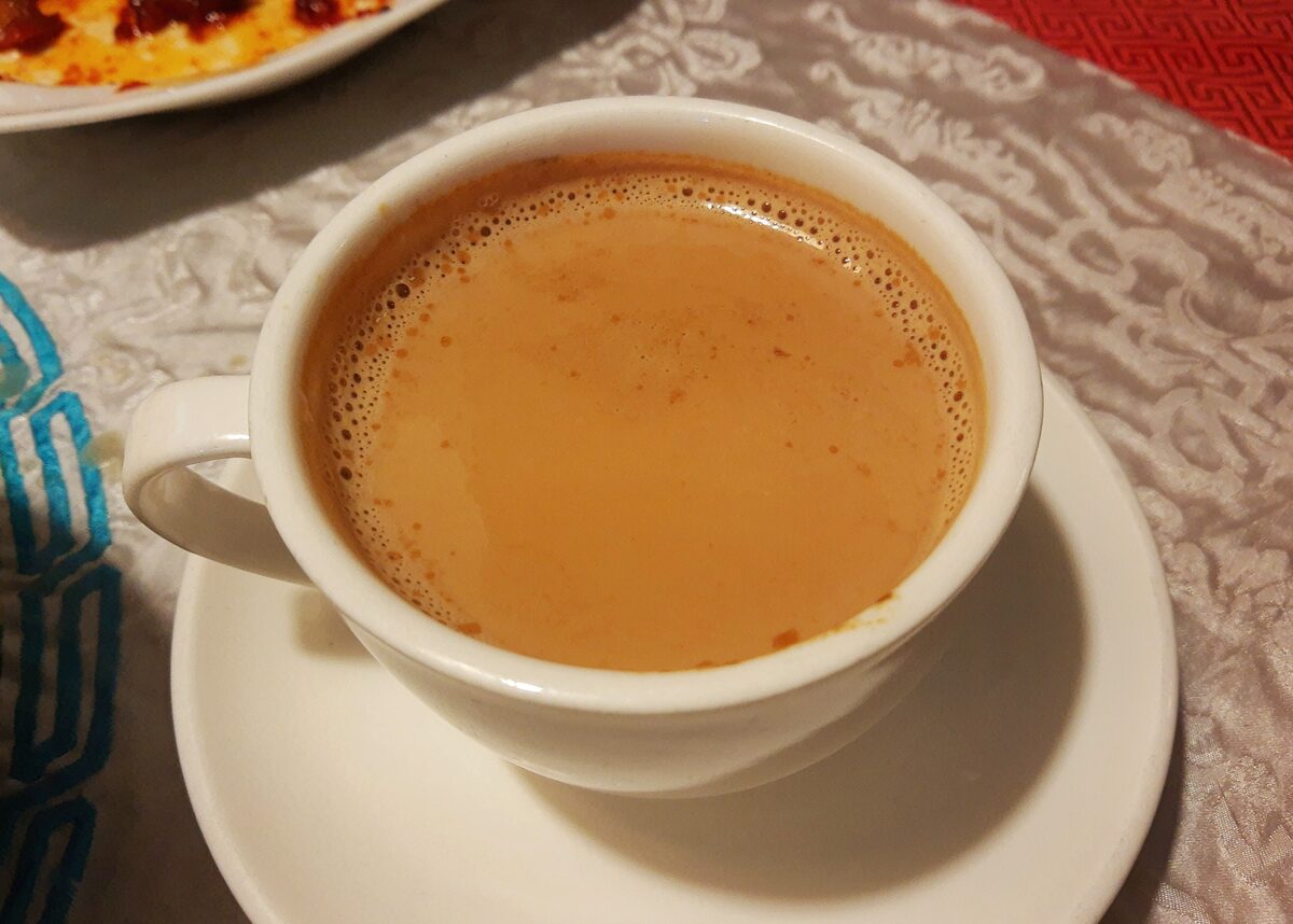 A white cup of frothy milk tea on a saucer, set on a silver brocade tablecloth