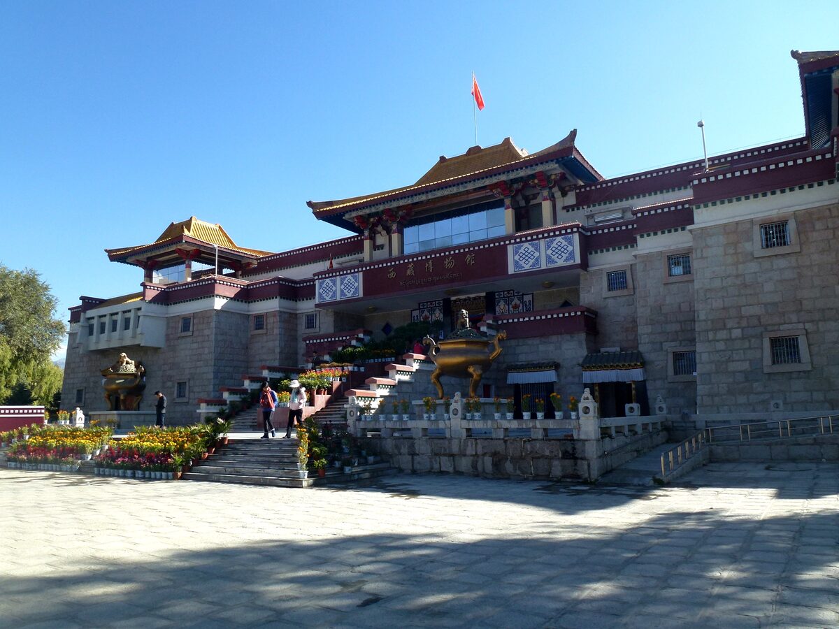 The entrance of the Tibet Museum, a stone-clad building with Tibetan-style red trim and golden roofs, with steps and flower beds in front