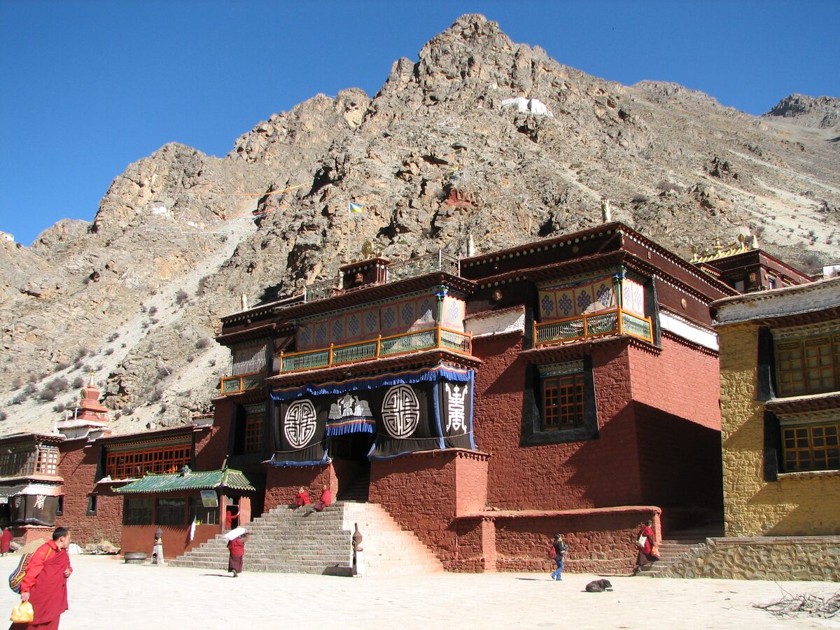 A red-walled Tsurphu Monastery building with a stone stairway, below a bare rocky mountain and blue sky
