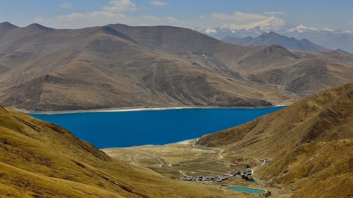 Deep turquoise Yamdrok Lake between brown mountain slopes, with a small village and fields at its edge