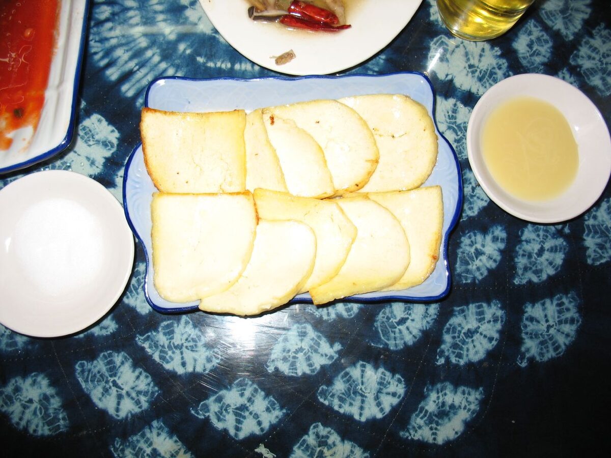 Plain slices of pale rubing goat cheese, the cheese itself before it is fried, on a blue tie-dye tablecloth with two small dishes of dip