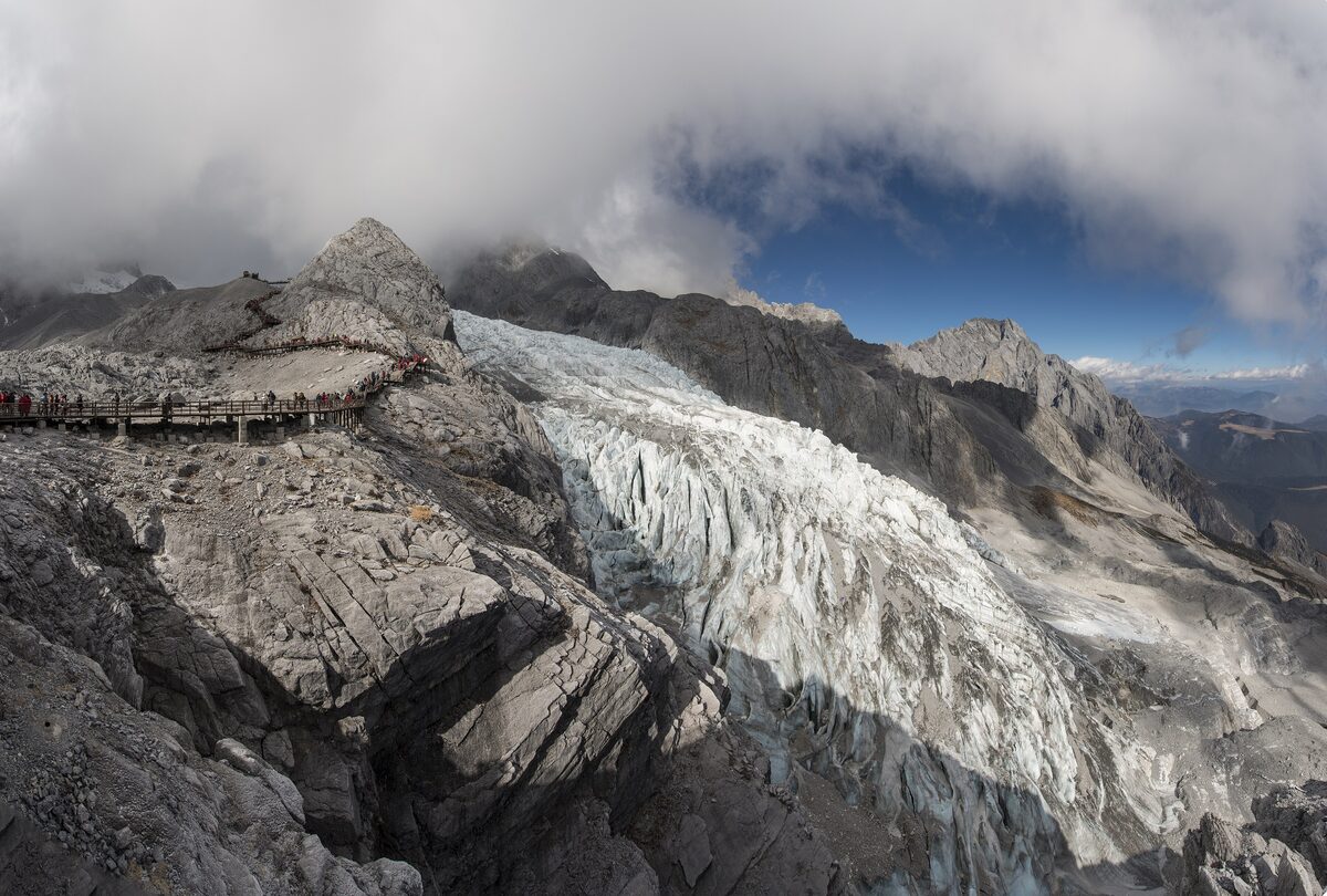 A glacier tongue of Jade Dragon Snow Mountain between grey rock ridges, with a boardwalk on the ridge at left and clouds opening above