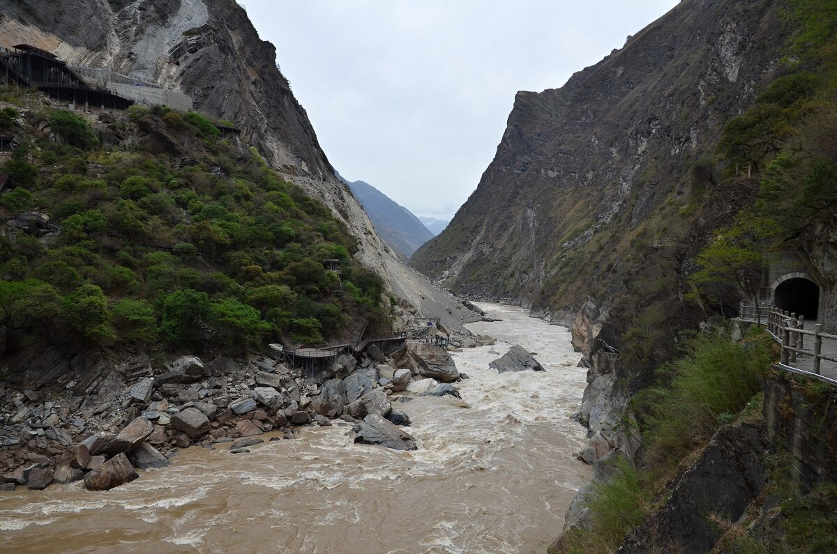 The brown Jinsha River running between steep rock walls in Tiger Leaping Gorge, with a tunnel mouth and a railed walkway on the cliff