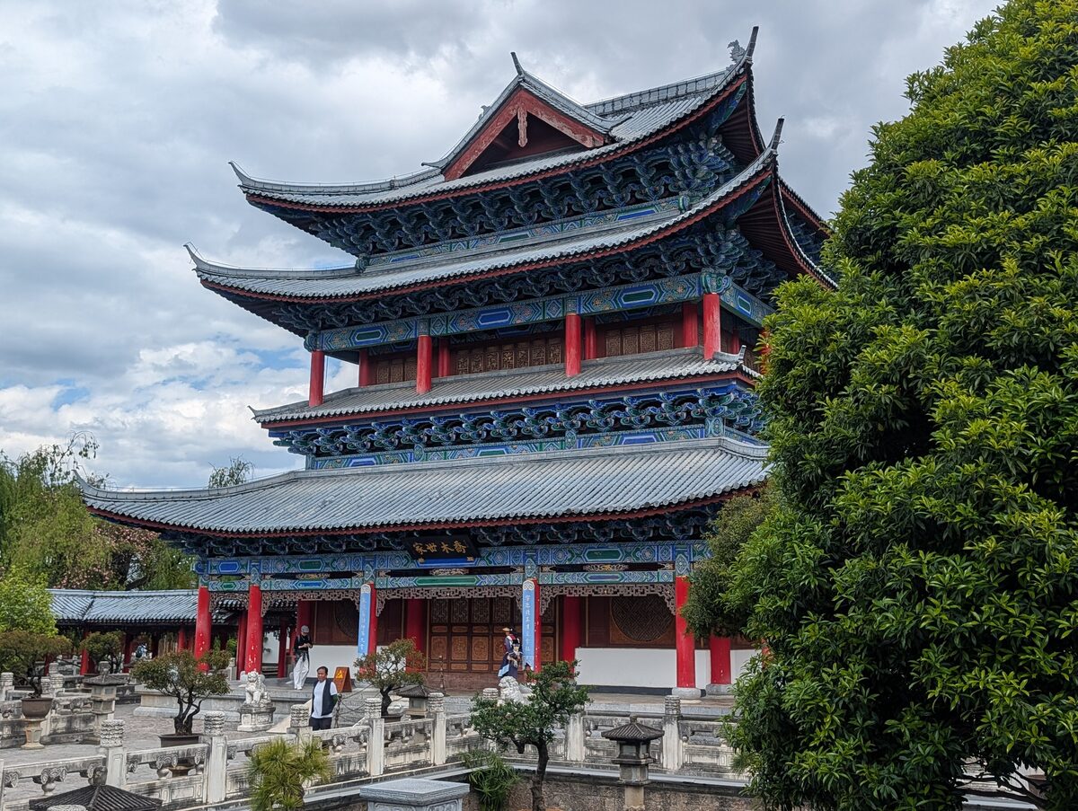 A tall hall with several tiled roofs and blue and red painted eaves in the Mu Palace compound, a leafy tree beside it
