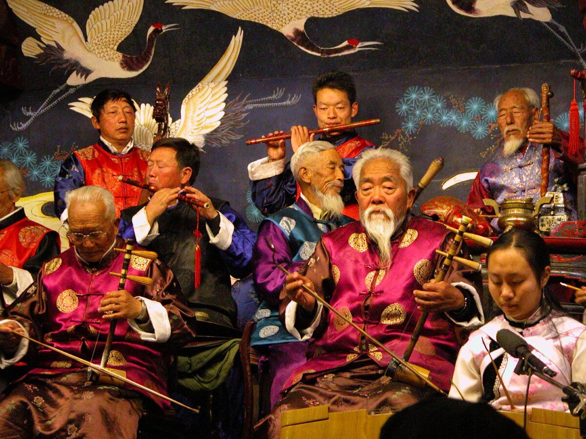 Elderly Naxi musicians in bright silk jackets playing string and wind instruments on a stage painted with cranes