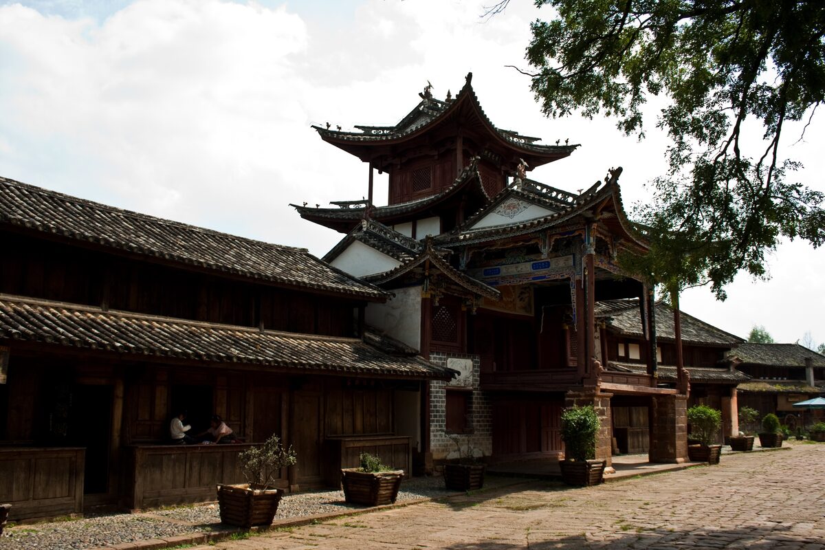 Old timber houses and a multi-roofed carved wooden building on a cobbled square in the old town of Shaxi, with a large tree overhead