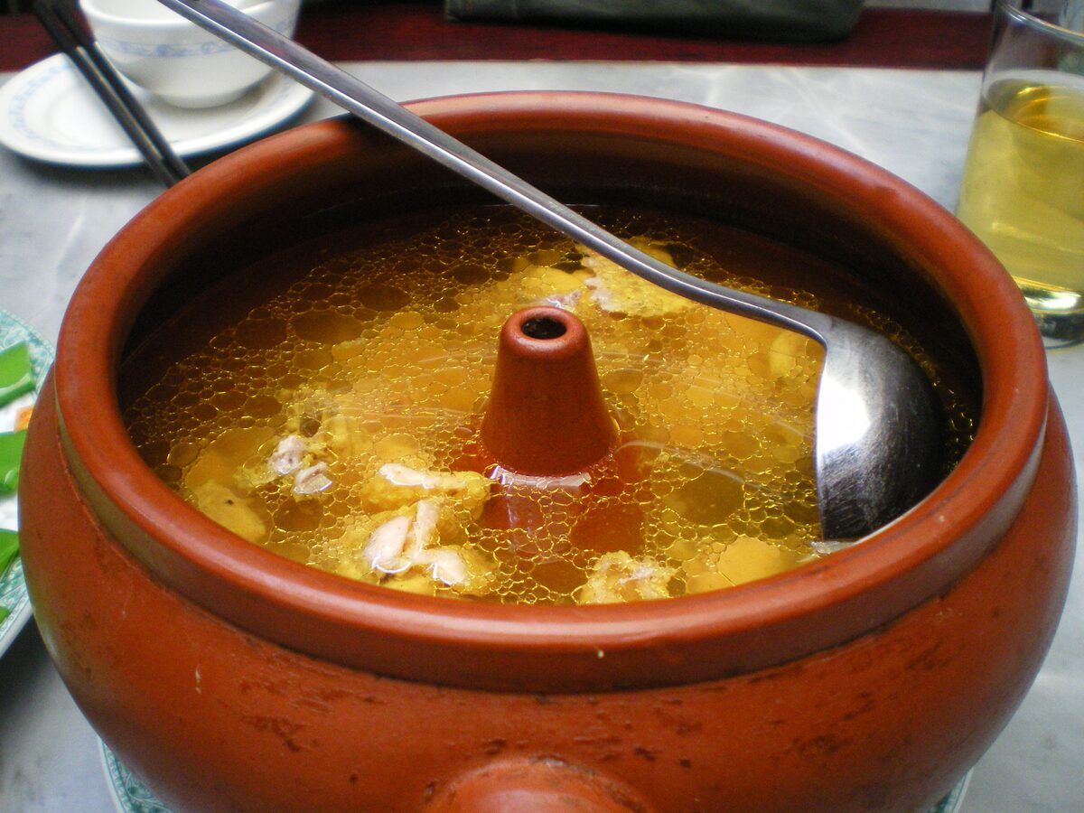 A clay steam pot of chicken soup with a central chimney and a steel ladle