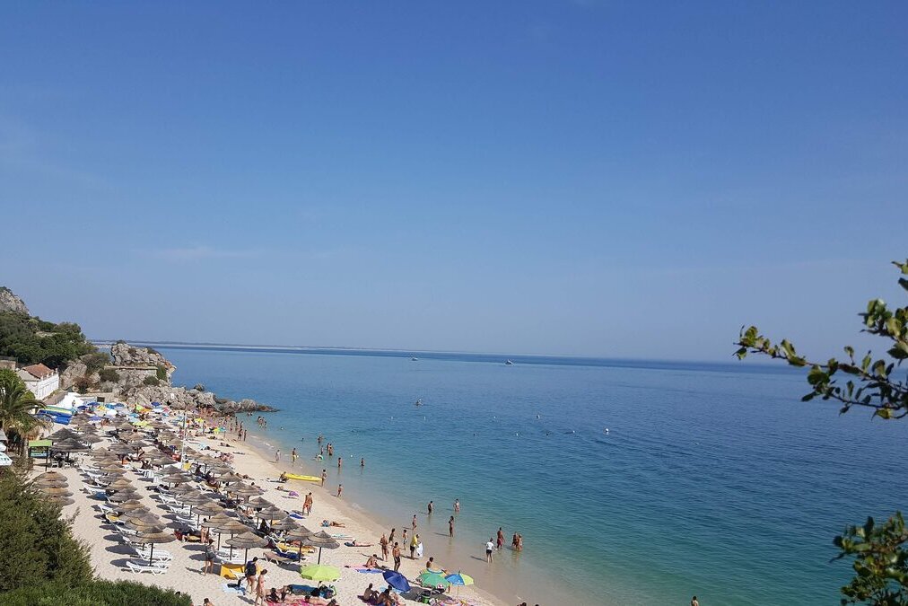 A sandy beach with rows of parasols under a limestone cliff on the Arrábida coast, the sea blue to the horizon