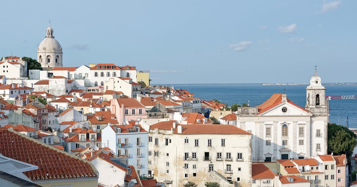 Red-roofed houses of Alfama tumbling down toward the Tagus from Miradouro de Santa Luzia, with the National Pantheon dome and the church of Santo Estêvão in view