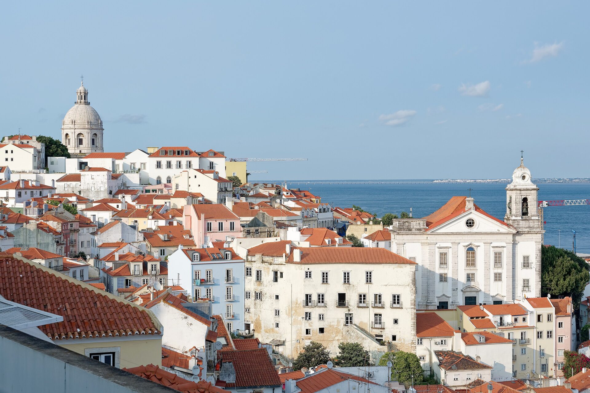 Red-roofed houses of Alfama tumbling down toward the Tagus from Miradouro de Santa Luzia, with the National Pantheon dome and the church of Santo Estêvão in view