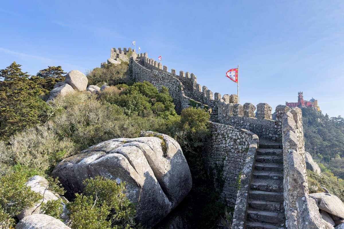 Stone steps climbing the Moorish Castle wall past a granite boulder, with a flag and the red tower of Pena Palace behind
