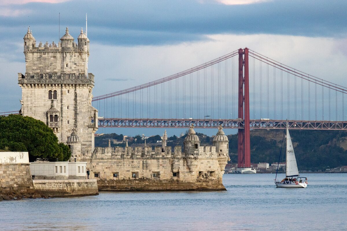 Belém Tower on the edge of the Tagus with the red 25 de Abril Bridge behind it and a sailboat passing