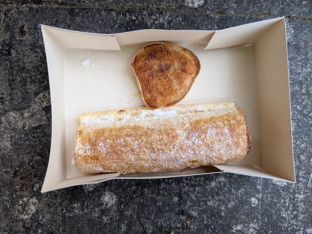 A travesseiro, a sugar-dusted puff pastry roll, next to a small round queijada in a paper tray