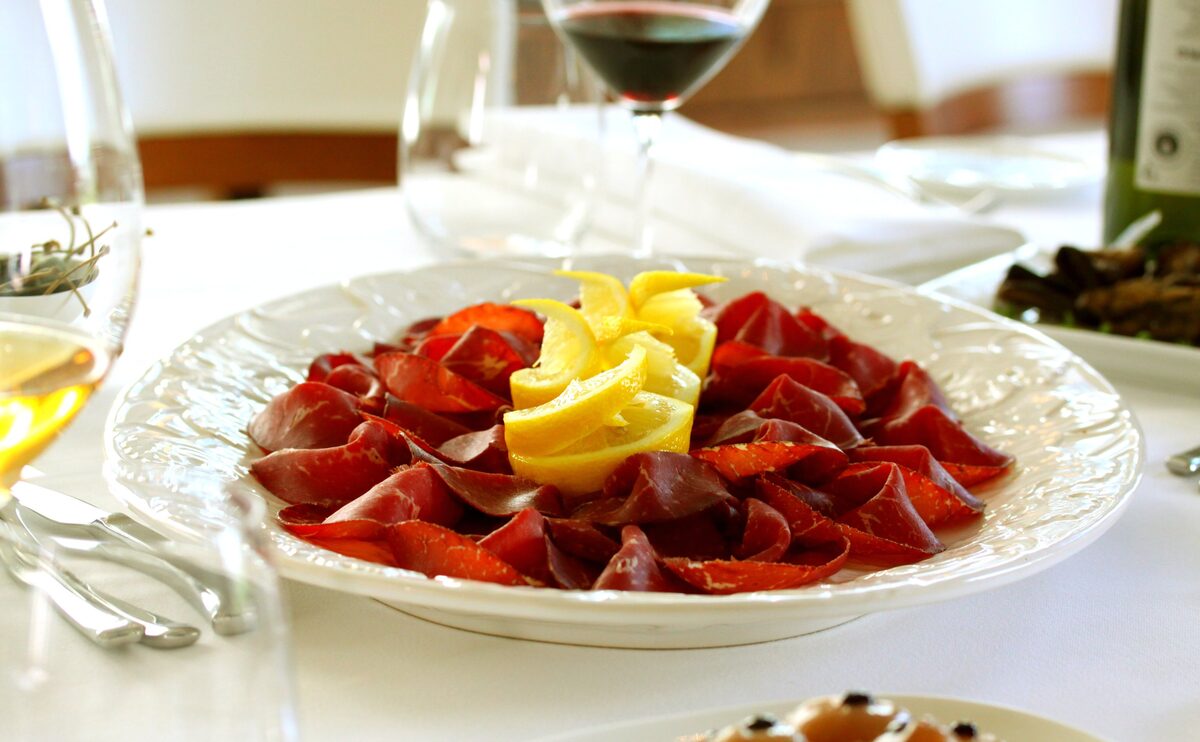 A white plate of thinly sliced bresaola arranged around a lemon twist, with a glass of red wine beside it