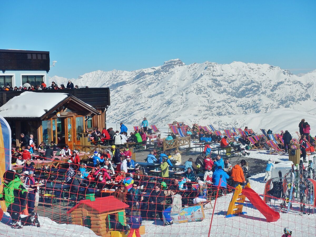 A busy sunny mountain terrace in Livigno with picnic tables, deck chairs and a children's slide, snowy peaks behind