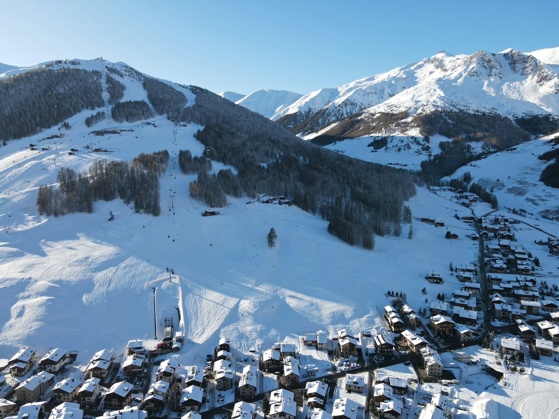Aerial view of the snowy slopes and chalet roofs of Livigno on a clear winter day, with a ridge of white peaks behind