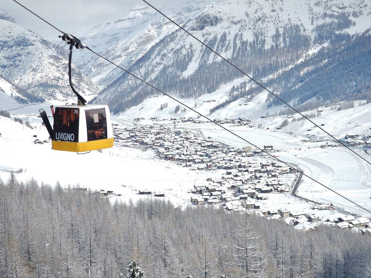 A yellow gondola car above the snowy village of Livigno, with larch trees on the slope below