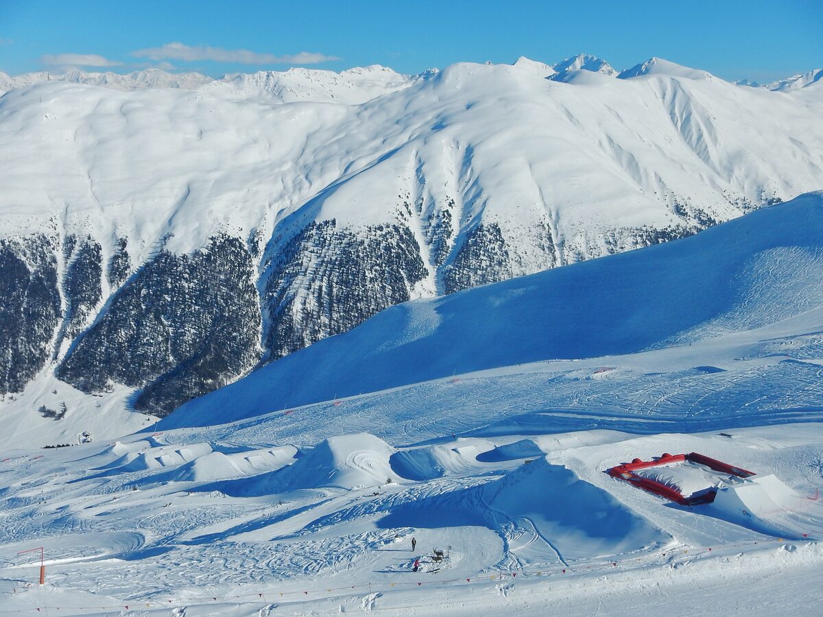 A snowpark with small jumps and a red airbag landing on the Carosello 3000 slopes, with snowy peaks behind