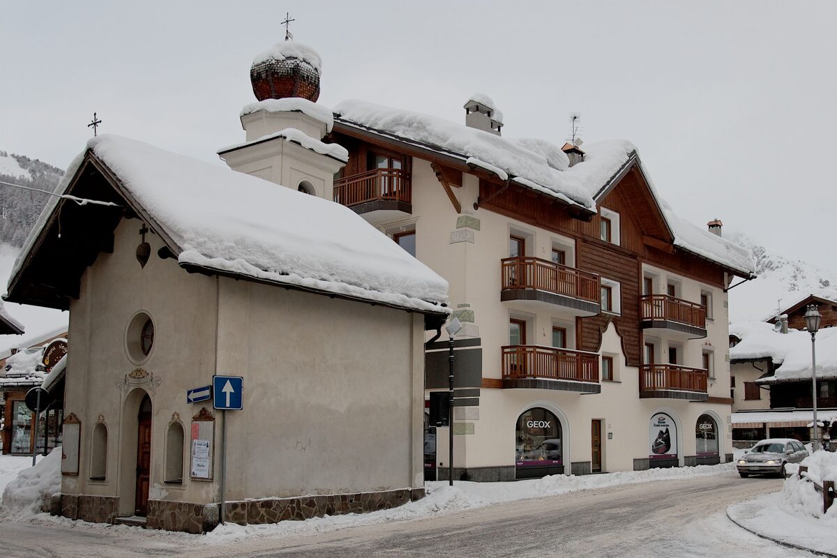 A snowy street corner in the Livigno center with a small chapel and a building with wooden balconies and shop windows