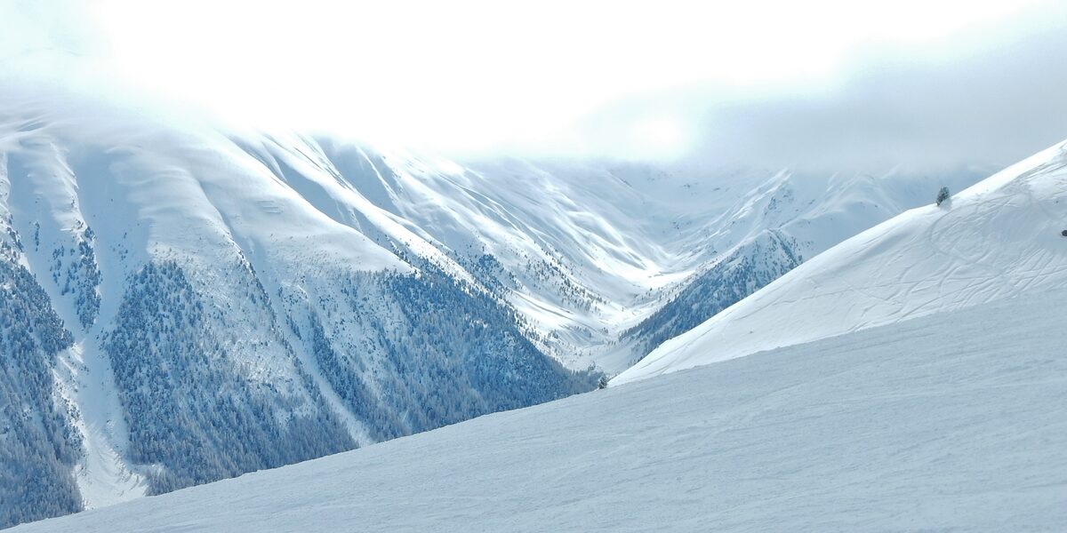 A snowy ridge in the Livigno ski area with mist over a deep valley and forested slopes falling away below
