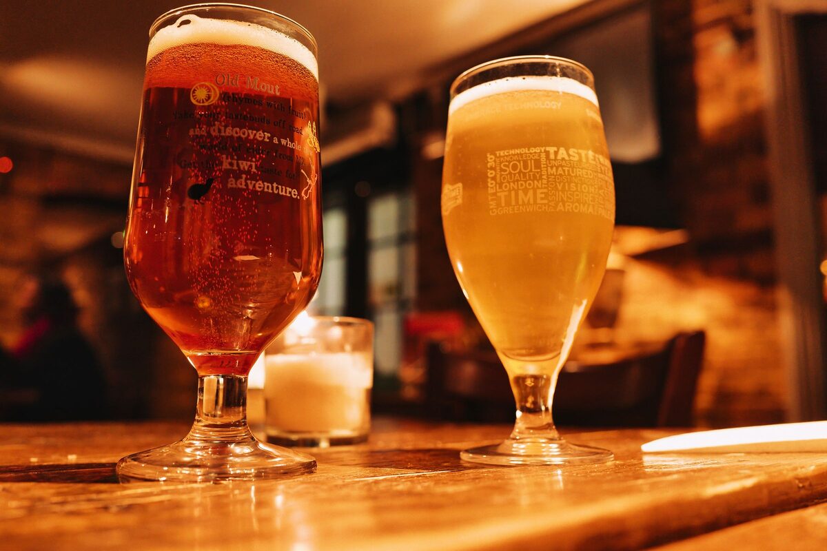 Two glasses of beer, one amber and one pale, on a wooden table in a warmly lit pub