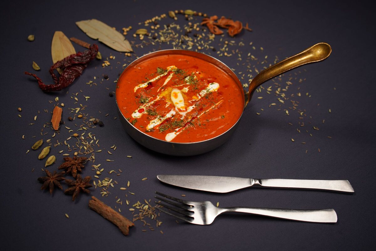 A pan of chicken tikka masala in an orange creamy sauce, with spices scattered around it