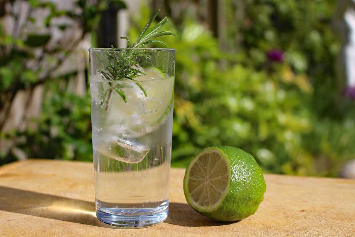 A gin and tonic with ice and rosemary next to a halved lime on a wooden table