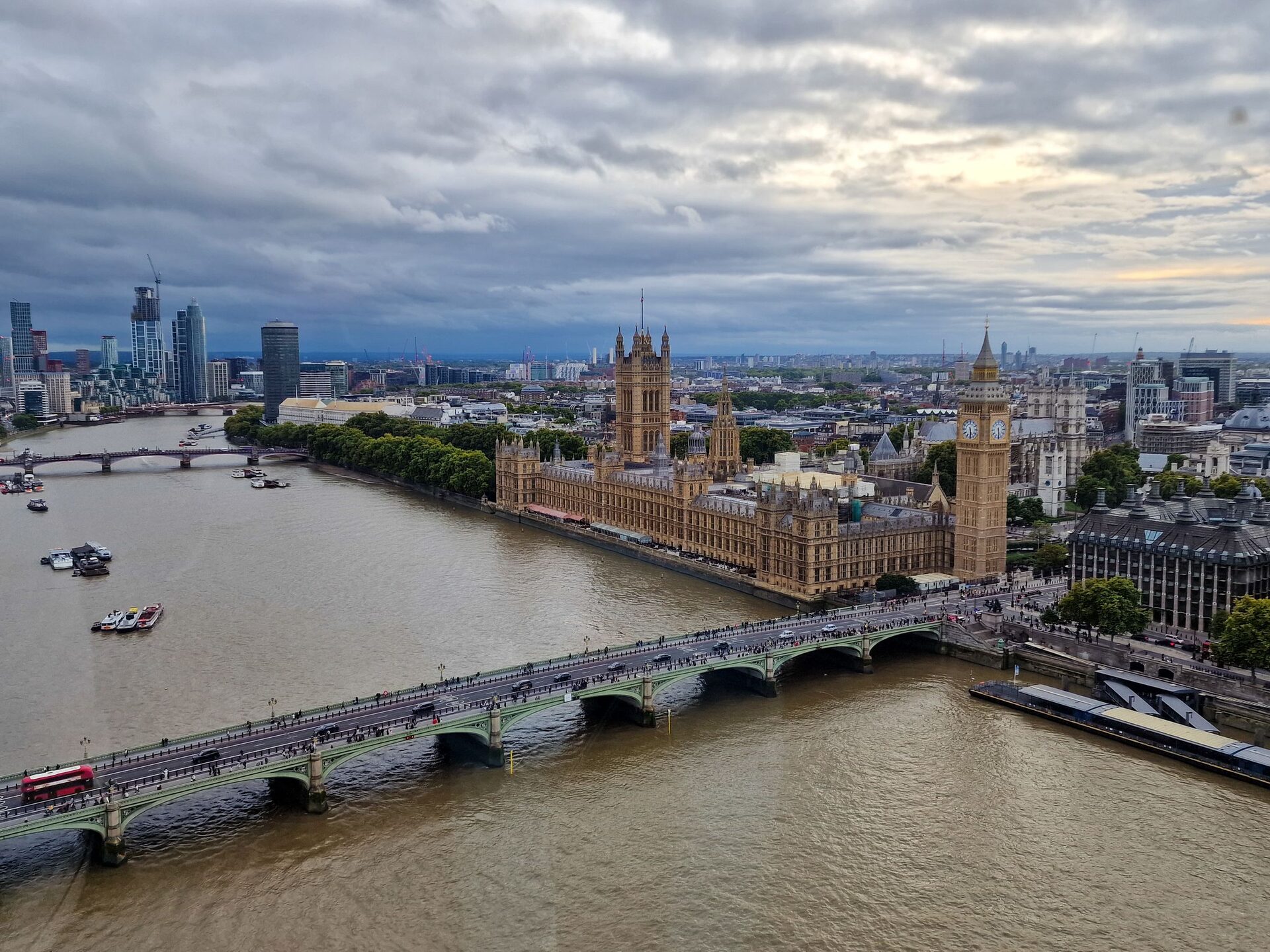 Aerial view of the Houses of Parliament and Big Ben beside the River Thames in London, with Westminster Bridge in front and the city spreading behind under a cloudy sky