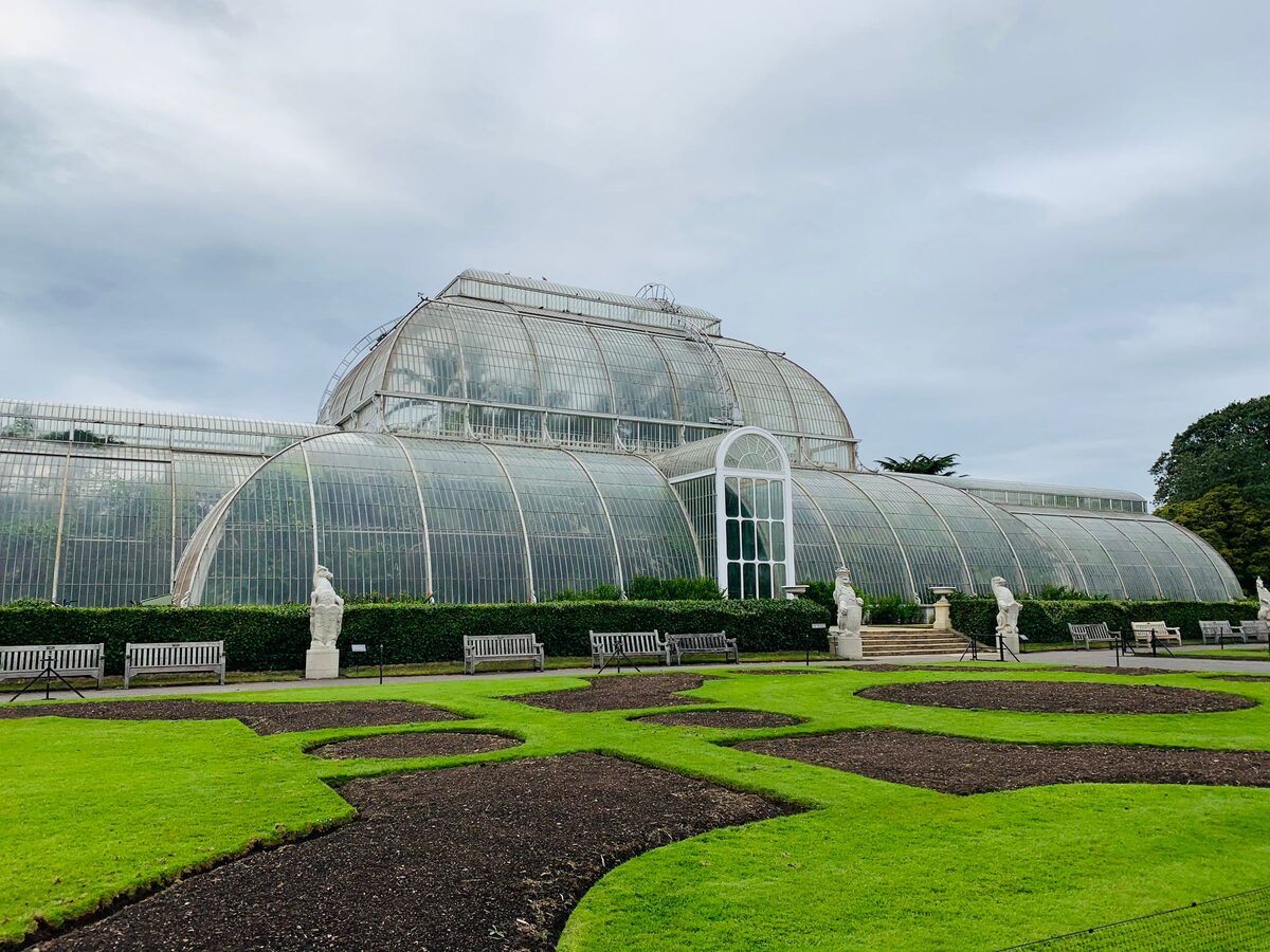 The Victorian Palm House at Kew Gardens, a curved glass and iron greenhouse, reflected in a pond