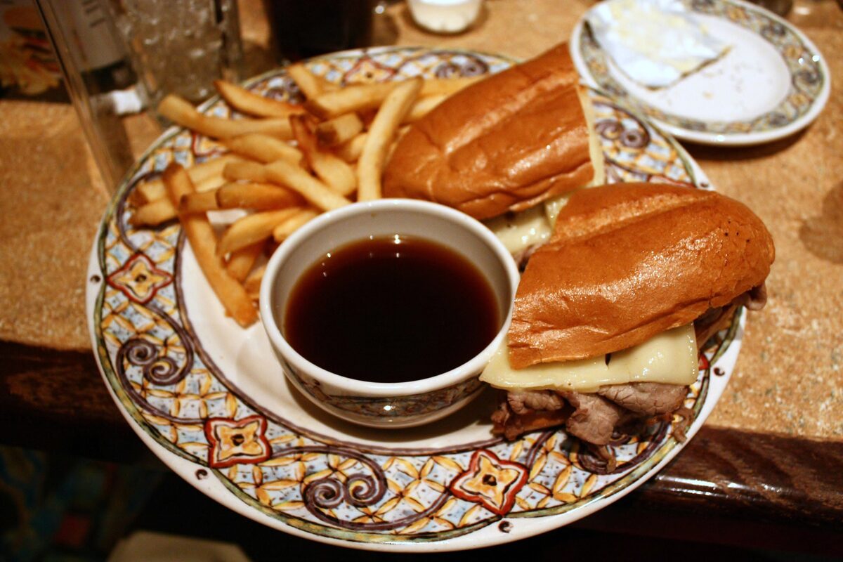 A French dip sandwich of sliced roast beef and cheese on a roll with a cup of au jus and fries