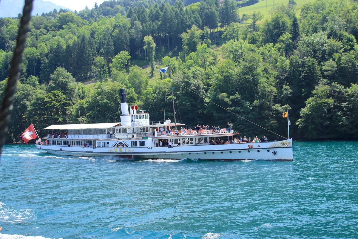 A white paddle steamer of the Lake Lucerne fleet with a Swiss flag, sailing on turquoise water below wooded slopes