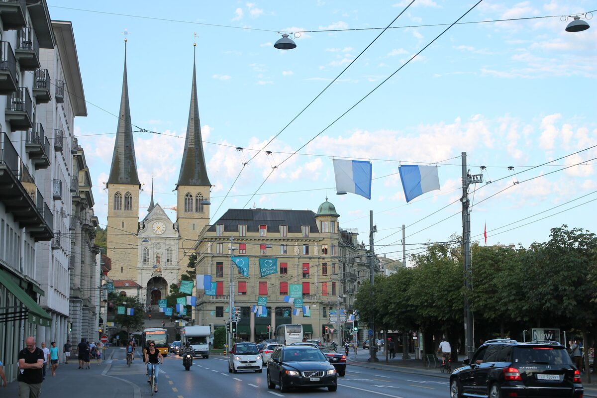 The twin spires of the Hofkirche St. Leodegar at the end of a busy street, with flags hanging above it