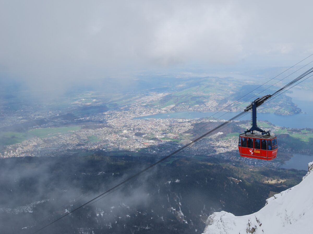 A red Pilatus gondola on its cable above the Lucerne basin, with the city and the lake far below