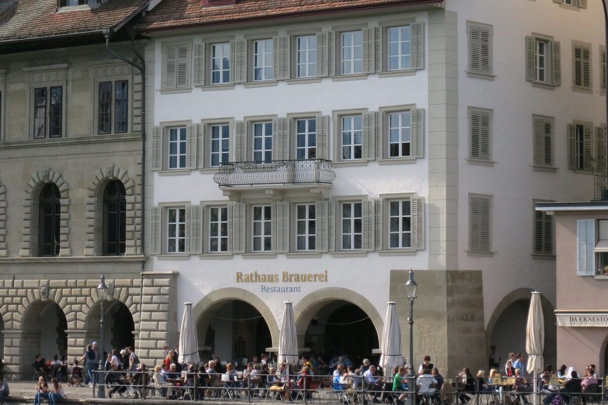The cream-coloured Rathaus Brauerei building on the Rathausquai, with its restaurant sign above the arches and guests on the riverside terrace