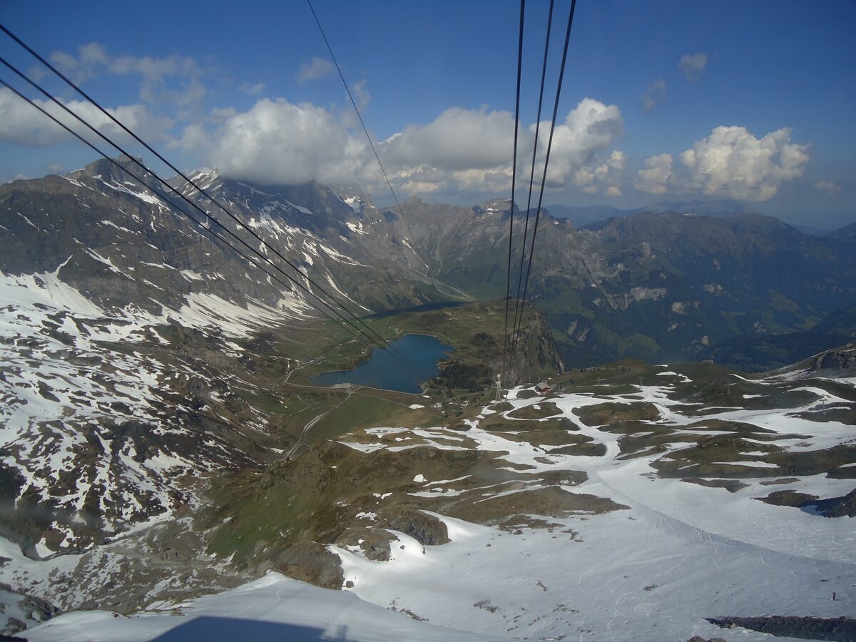 The Titlis cable car cables high above a small mountain lake and snow patches, with clouds over the ridges