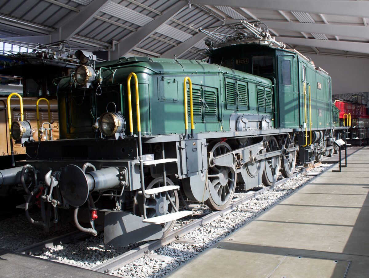 A green Swiss electric locomotive on display in a museum hall, with its pantograph raised
