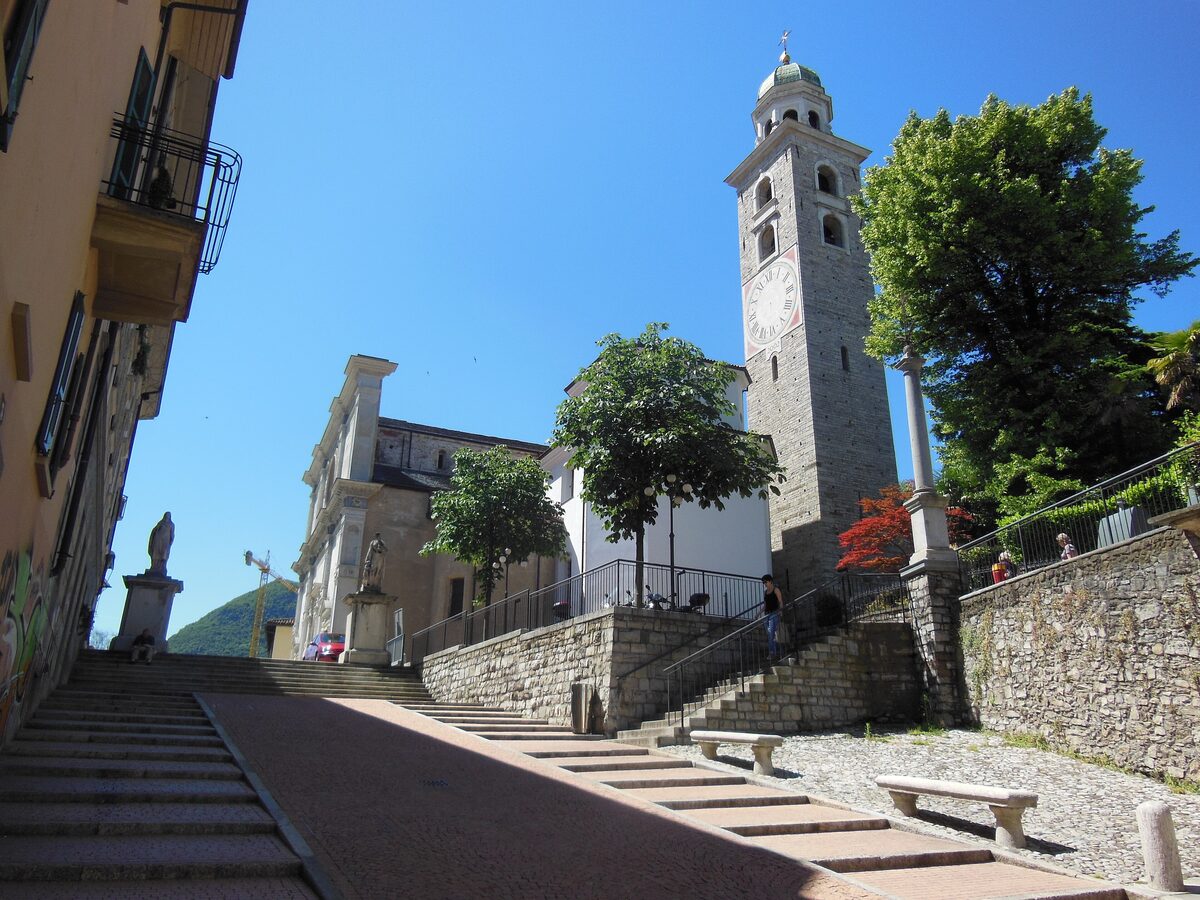 The bell tower of Lugano Cathedral rising above a steep stone lane on a clear day