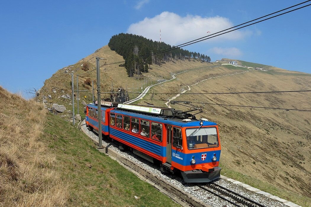 A blue and orange Monte Generoso rack train climbing the bare upper slope toward the summit