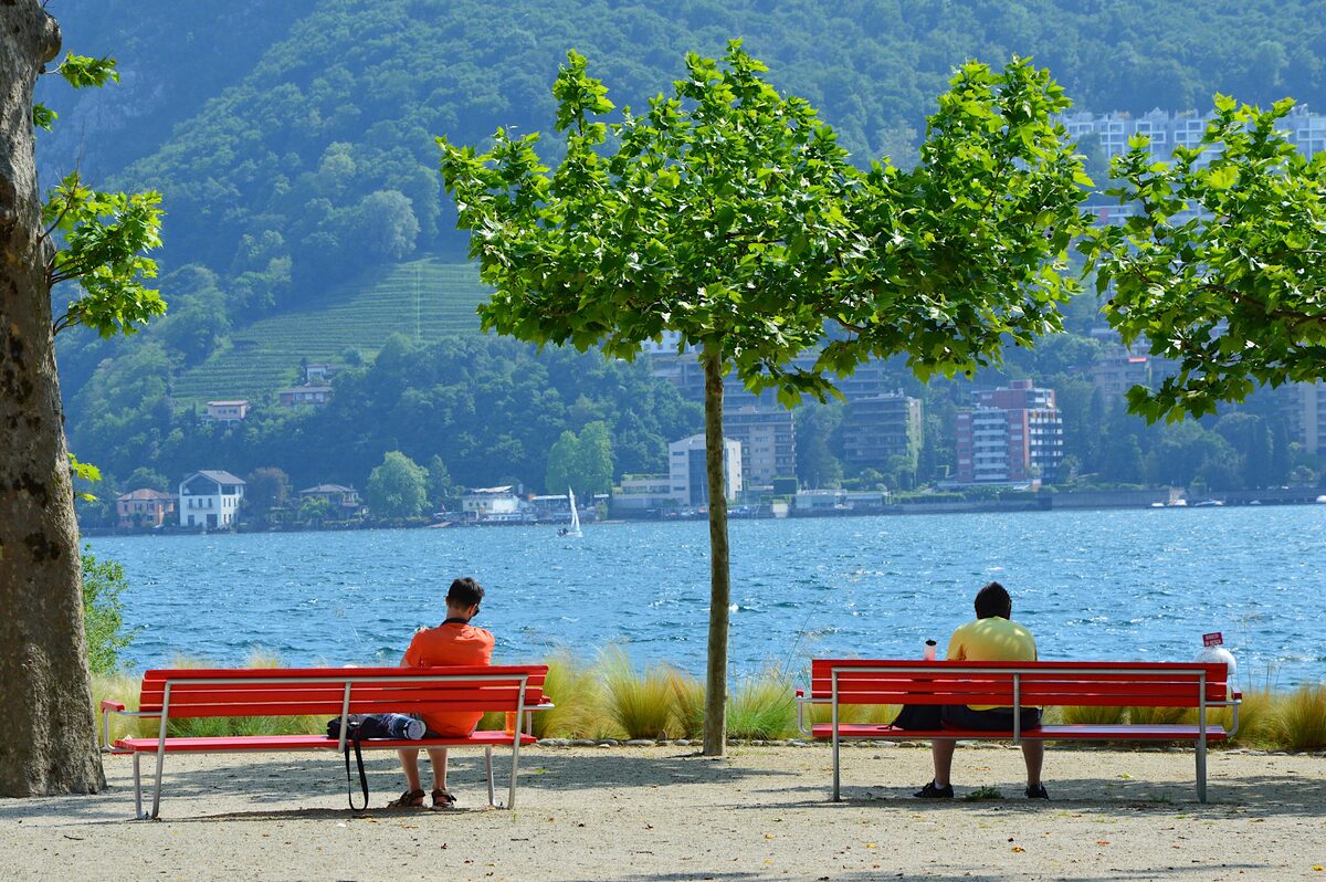 Red benches facing the lake on the Lugano waterfront, with trees and a green hillside across the water